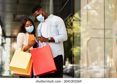 Loyalty Program, Cashback Concept. Portrait Of Happy Black Couple In Surgical Face Masks Using Smartphone Checking Bank Account After Successful Shopping In Mall During Covid Pandemic, Free Copy Space