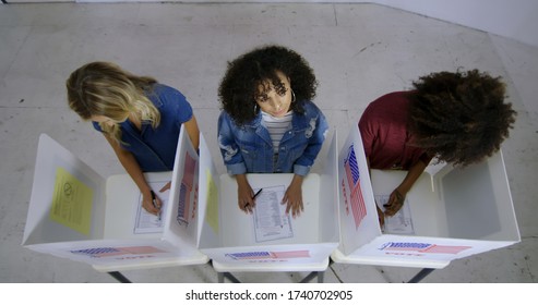 LS Wide overhead view three young women with HispanicLatina woman looking up and thinking while completing ballots at voting booths in polling station.