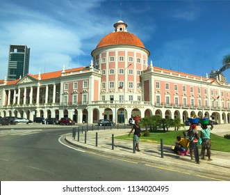 LUANDA, ANGOLA - MARCH 29 2018: Vendors Carrying Baskets On Their Heads In Front Of Beautiful  National Bank Of Angola Building In Downtown Luanda