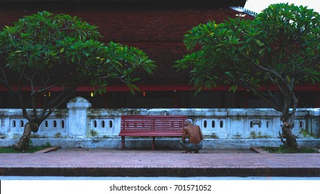 LUANG PRABANG, AUGUST 2017: An Old Man Squatting Down In Front Of A Red Bench Outside Wat Mai