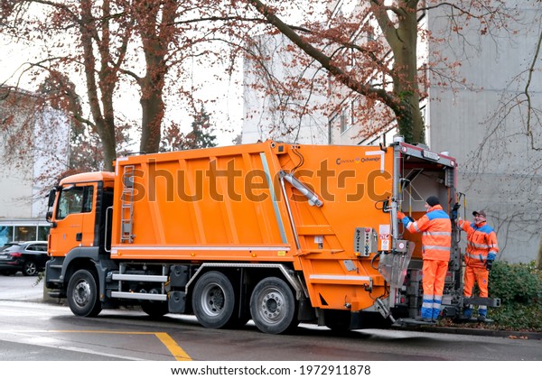 LUCERNE, SWITZERLAND - January 2021: orange cargo\
garbage truck driving through city streets, collecting garbage from\
bins, waste recycling concept, traffic safety regulation concept on\
streets