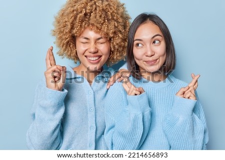 Luck and expectations concept. Happy multiethnic women cross fingers make wish have desire dreams come true dressed in knitted jumpers stand closely to each other isolated over blue background.