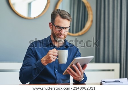 Luckily the coffee is tasting good today. Shot of a focused middle aged businessman browsing on a digital tablet while enjoying a cup of coffee at home during the day.