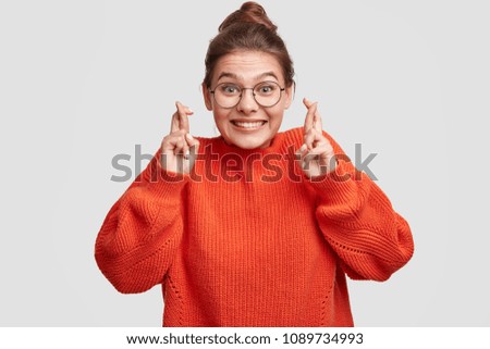 Lucky beautiful Caucasian female crosses fingers , hopes all wishes come true, wears oversized red sweater and spectacles, isolated over white background. People, body language and happiness