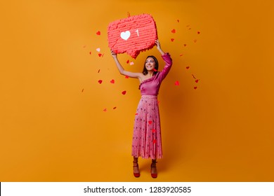 Lucky, Charming Woman Of 23 Years Holds Sign In Form Of Like From Instagram And Poses In Full Growth On Orange Background With Confetti