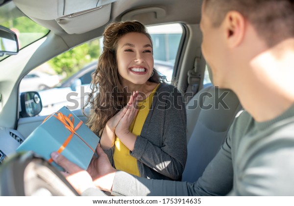 Lucky day. Happy\
shining woman with white-toothed smile looking at man with gift box\
in hands, sitting in car