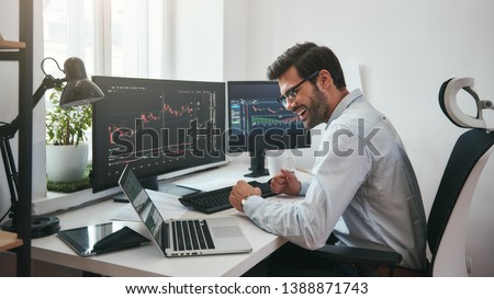 Lucky day. Happy young businessman or trader in formalwear and eyeglasses using laptop and smiling while sitting in his modern office