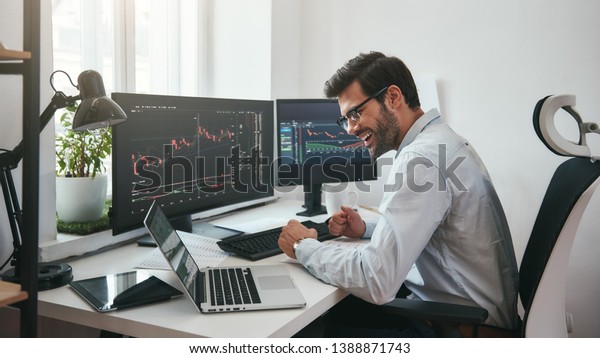 Lucky day. Happy young businessman or trader in\
formalwear and eyeglasses using laptop and smiling while sitting in\
his modern office