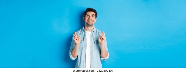 Lucky guy praying and making wish with crossed fingers, looking up with pleading face, standing against blue background.