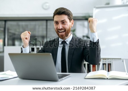 Lucky happy Caucasian business man, sales manager, executive, sits in front of a laptop screen at his desk, in a modern office, insanely happy about a great deal, big profit, gesturing with his fists