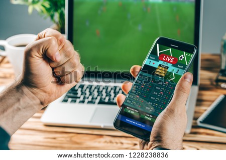 Lucky man celebrating victory after making bets using gambling mobile application on his phone. Football match online broadcast on laptop screen on the background.