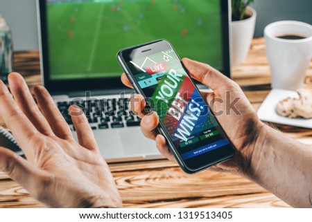 Lucky man celebrating victory after making bets using gambling mobile application on his phone. Football match online broadcast on laptop screen on the background. 