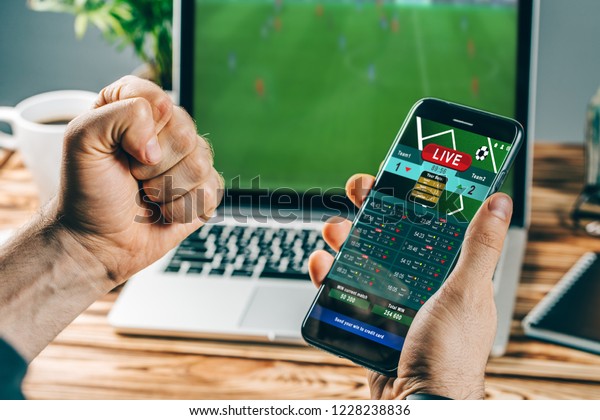 Lucky man celebrating victory\
after making bets using gambling mobile application on his phone.\
Football match online broadcast on laptop screen on the\
background.