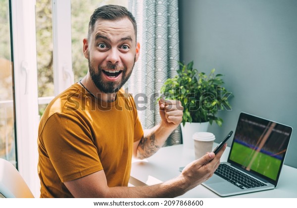 Lucky man\
celebrating victory after making bets at bookmaker\'s website.\
Football fan watching play live broadcast on his laptop computer\
and  cheering for his favourite\
team.