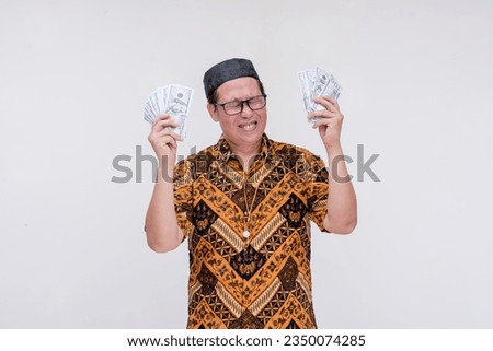 A lucky middle aged man celebrates, holding a lot of cash in his hand he won or earned. Wearing a batik shirt and songkok skull cap. Isolated on a white background.