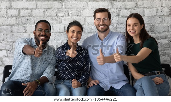 Lucky ones. Portrait of four happy satisfied\
multiethnic persons sitting on row of chairs in office looking at\
camera showing thumbs up being hired recruited getting good\
positions in corporate\
staff