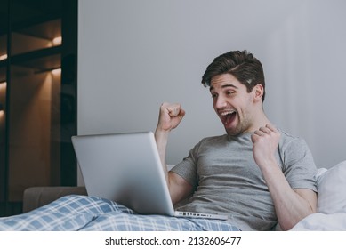 Lucky Overjoyed Excited Young Man In Pajamas Grey T-shirt Sit In Bed Hold Use Work On Laptop Pc Computer Clench Fist Rest Relax At Home Indoors Bedroom Good Mood Sleeping Night Morning Bedtime Concept