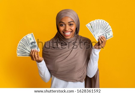 Lucky Winner. Overjoyed Black Muslim Girl in Hijab Holding Lots Of Dollar Cash In Both Hands, Standing Over Yellow Background With Free Space