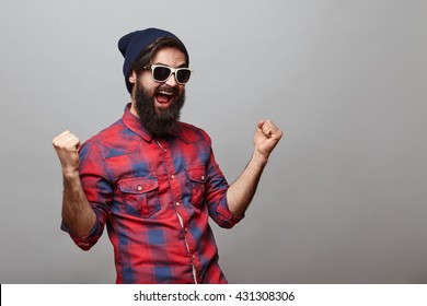 Lucky young hipster man isolated over grey background. Bearded man wearing glasses and hat acts like he won something with copy space - Powered by Shutterstock