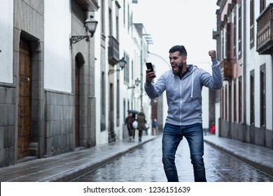 Lucky young man screaming with fist up holding cellphone on rainy street in the city. Full beard hipster on grey hoodie with euphoric expression. Online bet winner, victory celebration concepts - Powered by Shutterstock