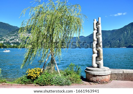 Lugano, Switzerland - A boardwalk on the shore of Lake Lugano, a tree and yellow flowers, next to a sculpture in the form of an anchor chain, on the back ground are alpine mountains, a blue sky.