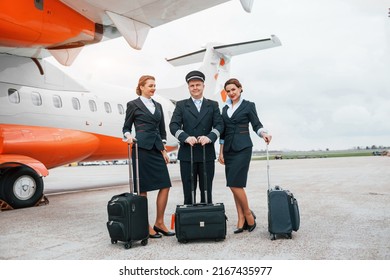 With Luggage. Aircraft Crew In Work Uniform Is Together Outdoors Near Plane.