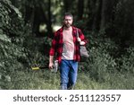 The Lumberjack holds axe with serious face on the forest. A lumberjack with a large ax examines the tree before felling. Serious lumberjack. Confident young bearded man holding a big axe outdoor.