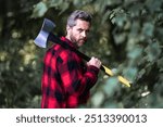 The Lumberjack holds axe with serious face on the forest. A lumberjack with a large ax examines the tree before felling. Serious lumberjack. Confident young bearded man holding a big axe outdoor.