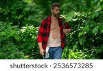 The Lumberjack holds axe with serious face on the forest. A lumberjack with a large ax examines the tree before felling. Serious lumberjack. Confident young bearded man holding a big axe outdoor.