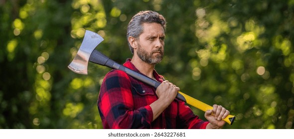 Lumberjack men in forest with axe. Lumberjack carries axe. Handsome man with axe. Lumberjack in woods with an ax. Lumberjack in shirt with heavy axe. Strong man work in forest. Focused expression