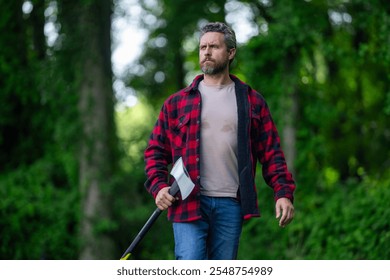 Lumberjack with serious face carries axe outdoor. Lumberjack worker in the forest with axe. Strong man lumberjack with an ax in a plaid shirt. Lumberjack or logger in forest.