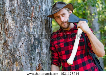Lumberjack worker standing in the forest with axe. Lumberjack holding the axe. Strong man lumberjack with an ax in a plaid shirt. Lumberjack with ax in his hands