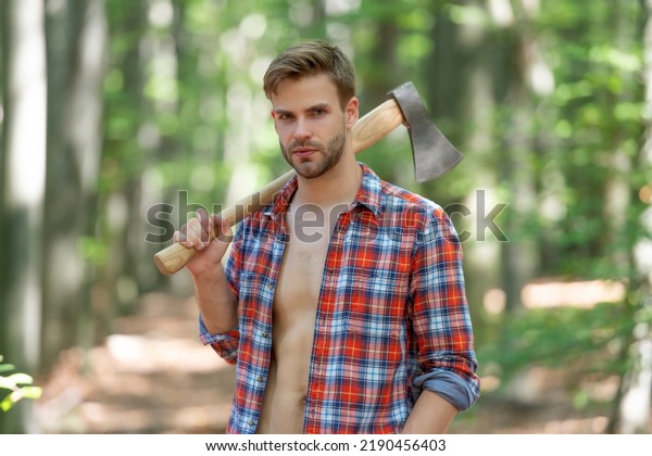 Lumbersexual guy in lumberjack shirt holding\
axe on shoulder forest\
background