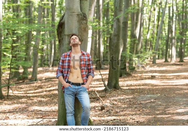 Lumbersexual man with closed eyes\
in unbuttoned lumberjack shirt forest background, copy\
space