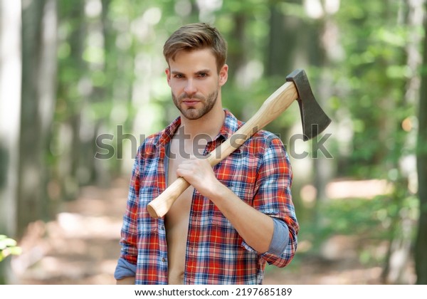 Lumbersexual man in lumberjack shirt holding\
axe on shoulder forest\
background