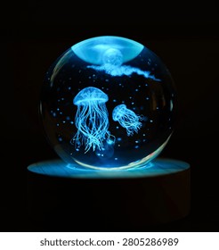 Luminous jellyfish gracefully float inside a clear glass sphere illuminating a dark room from a wooden base