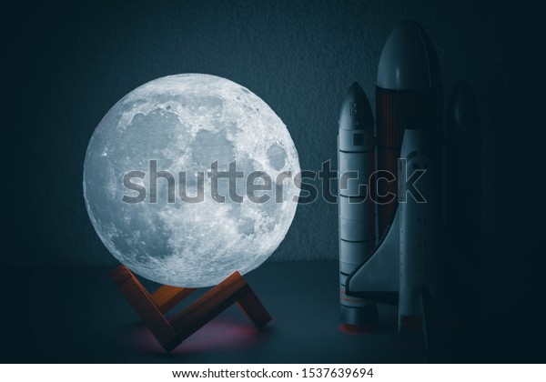 A luminous moon\
lamp next to a toy in the form of a rocket, all objects on a light\
wooden background.
