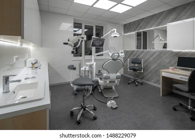 Luminous room in a dental clinic with light walls and dark gray floor. There is a workplace with a dental chair and equipment, table with a computer, zone with a sink, microscope, wide mirror, window. - Shutterstock ID 1488229079