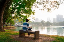 
Lumpini Park, Bangkok Thailand, A Young Asian Man Sitting In A Park Near The Pond With Views Of The City Skyline