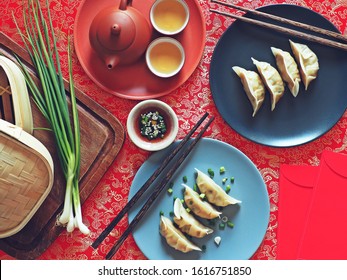 Lunar New Year Lucky Food Chinese Steamed Dumplings On Red Fabric Background With Chopsticks, Red Envelopes, Bamboo Basket, Green Onions, Soy Sauce, Tea Set. Traditional Dish Eaten On Chinese New Year