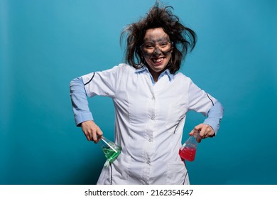 Lunatic biochemistry specialist grinning creepy while holding Erlenmeyer flasks filled with liquid substances. Insane looking mad chemist with dirty face and messy hair acting goofy on blue background