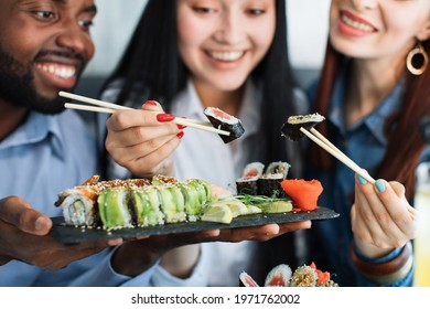 Lunch at a Asian restaurant. Close up shot of happy grinning multiethnic friends, black man and two pretty girls, eating sushi rolls with chinese sticks. Focus on plate with rolls and sticks - Powered by Shutterstock