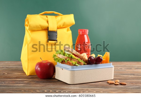 Lunch box with appetizing food and bag on\
wooden table against chalkboard\
background