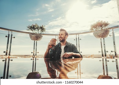 Lunch break. Bearded man holding coffee and smiling. He is sitting on the balcony of outdoor restaurant