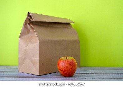 Lunch Packed In A Brown Paper Bag With Red Apple Isolated On Green Background And Wooden Desk. Healthy Food Concept. No People. Copy Space