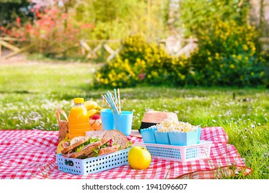 Lunch in the park on the green grass. Summer sunny day and picnic basket. Popcorn and sandwiches for a snack outdoors in nature. in bright plastic dishes on a checkered tablecloth . Copy space - Powered by Shutterstock