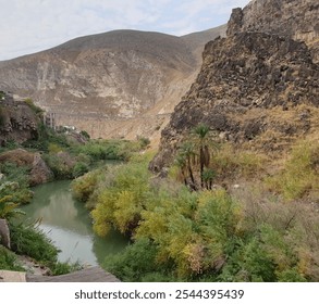 a lush, green river valley nestled between rugged, arid hills. The landscape features a mix of vegetation near the water and dry, rocky terrain on the surrounding cliffs. - Powered by Shutterstock