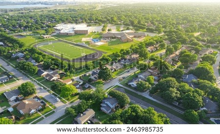 Lush green suburban neighborhood row of single-family houses, tall trees, spot complex football field, tennis courts, playground of middle elementary school in Dallas Fort Worth suburbs, aerial. USA