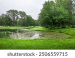 Lush greenery surrounds a peaceful pond, reflecting the overcast sky while soft ripples dance on the water