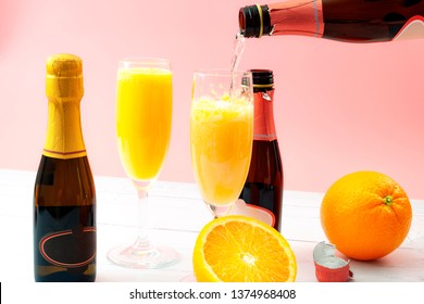 Lush Mix Drinks, Summer Alcoholic Beverages And Refreshing Brunch Mimosa Cocktail Concept With Orange Fruits And Champagne Pouring From A Bottle Of Bubbly Isolated On Wooden Table And Pink Background